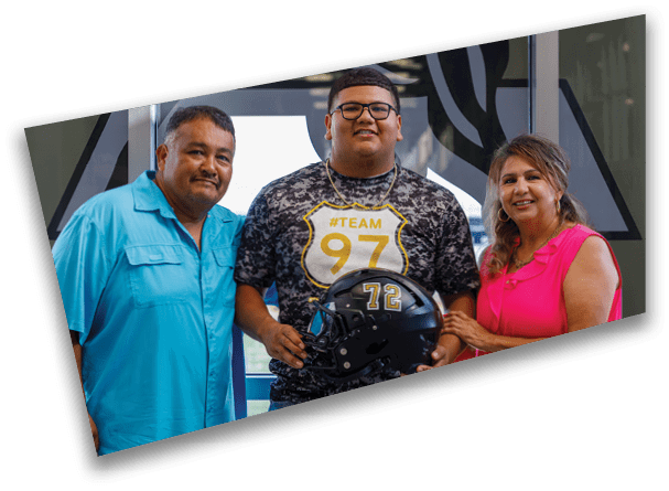 Three people stand closely together indoors with one person holding a football helmet and wearing a camouflage shirt with the text “#TEAM 97." Description generated by AI
