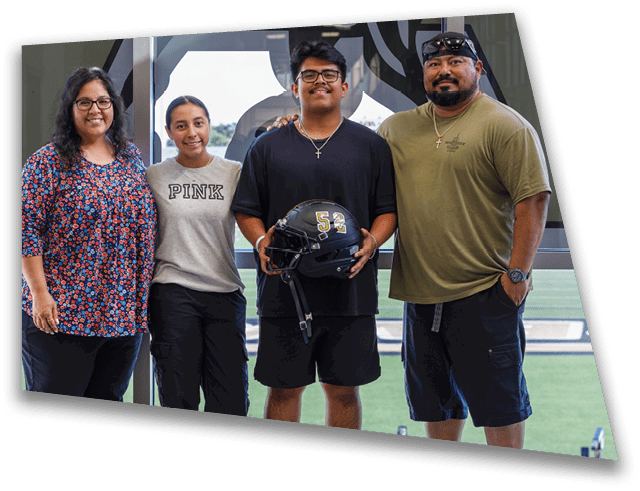Four adults stand closely together indoors near a window with a sports field visible outside, with one person holding a football helmet. Description generated by AI