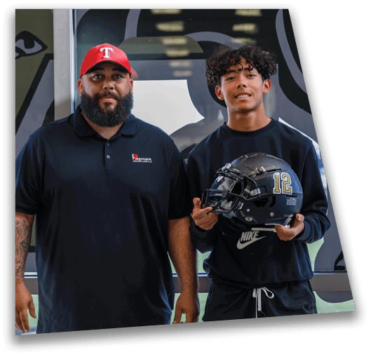 A person wearing a red baseball cap and black polo shirt stands next to another person holding a black football helmet in a sports setting. Description generated by AI