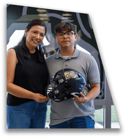 Two people stand indoors smiling while holding a black football helmet with the number 60 on it. Description generated by AI