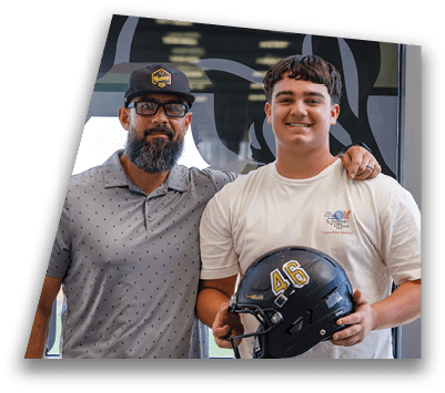 Two adults stand side by side indoors, one wearing glasses and a patterned polo shirt and the other holding a black football helmet. Description generated by AI