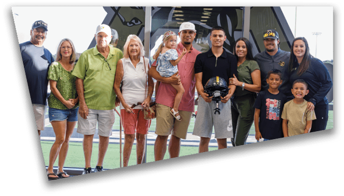A multigenerational group of people standing together outdoors on a sports field with some holding children and one person holding a football helmet. Description generated by AI