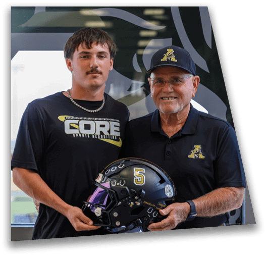 Two people stand indoors smiling while holding a black football helmet with the number five on it, both wearing dark shirts with matching logos. Description generated by AI