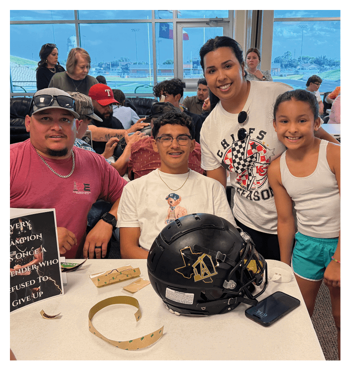 A group of people are gathered indoors with one person wearing glasses and a white t-shirt sitting in front of a black helmet while another person stands beside them wearing a white Chiefs season t-shirt and smiling. Description generated by AI