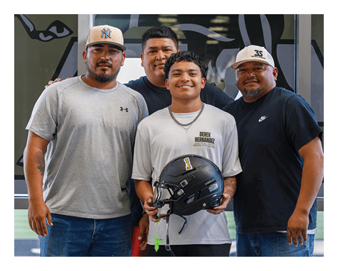 Four people stand closely together smiling indoors with one person holding a black football helmet. Description generated by AI
