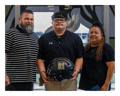 Three people stand indoors with one person holding a black football helmet with the number 21 on it, all appearing calm and posing for the photo. Description generated by AI