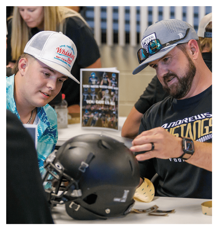 Two adults wearing casual clothing and hats are sitting at a table examining a black football helmet. Description generated by AI