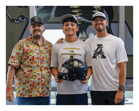 Three people stand side by side with one person in the center holding a black football helmet with the number eight, wearing a light-colored shirt that says “Andrews vs Everybody" above a large letter A. Description generated by AI