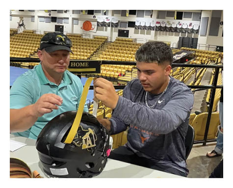 Two people are sitting at a table in a stadium setting, working together to attach a yellow strip to a black football helmet with a logo. Description generated by AI