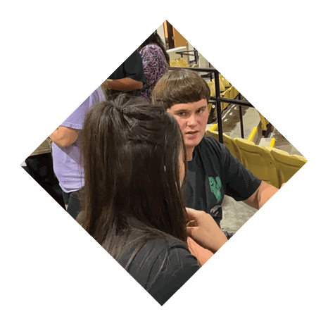 Two people are engaged in conversation near yellow stadium seats, with one holding a black and gold sports helmet. Description generated by AI