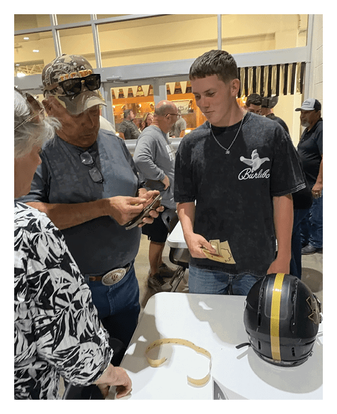 Three adults stand around a table with a black and gold helmet on it, one person holding cash and another using a smartphone, in an indoor setting with other people in the background. Description generated by AI
