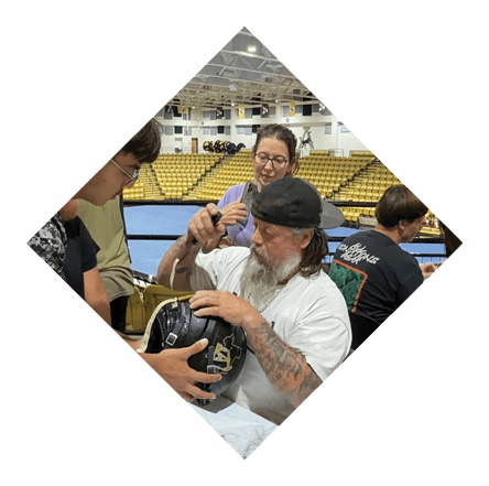 A person with a beard and tattoos wearing a backward cap is signing a helmet while others sit nearby in a gymnasium with yellow bleachers. Description generated by AI