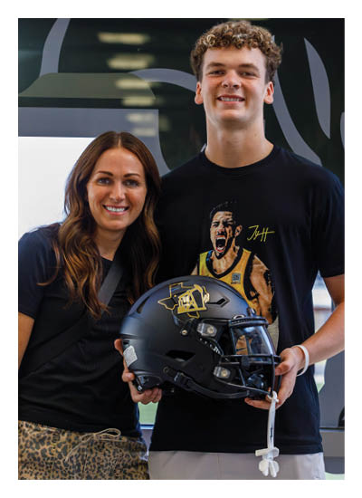 A person with long wavy hair wearing a black shirt and leopard print pants is smiling and holding a black helmet while standing indoors near a window. Description generated by AI