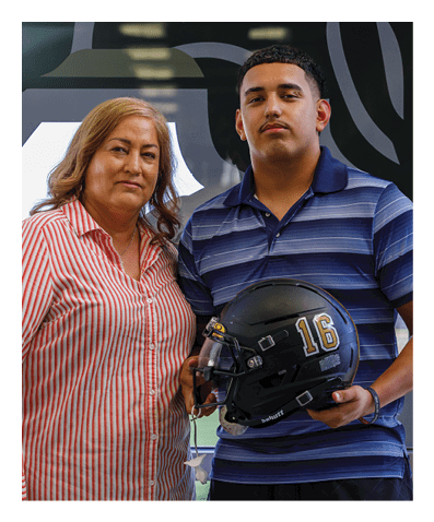 Two people stand closely together indoors, one wearing a striped shirt and the other holding a black helmet with the number 16 on it while dressed in a blue striped polo shirt. Description generated by AI