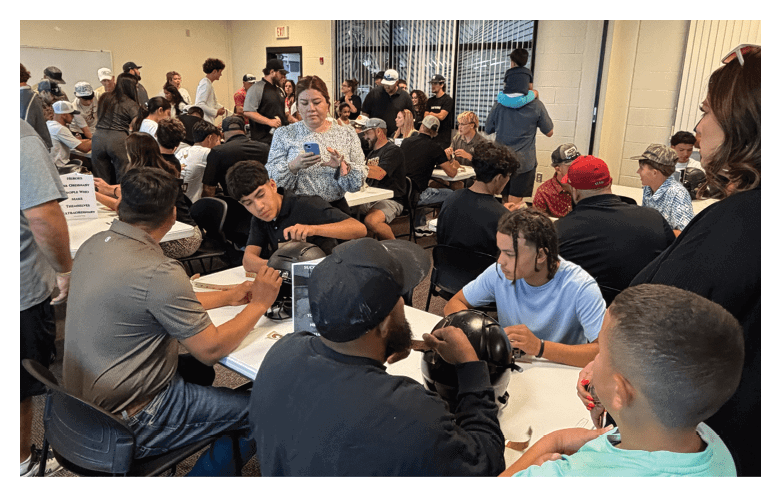 A large group of people seated closely together in a room appear to be engaged in a meeting or workshop, with some writing or using electronic devices. Description generated by AI