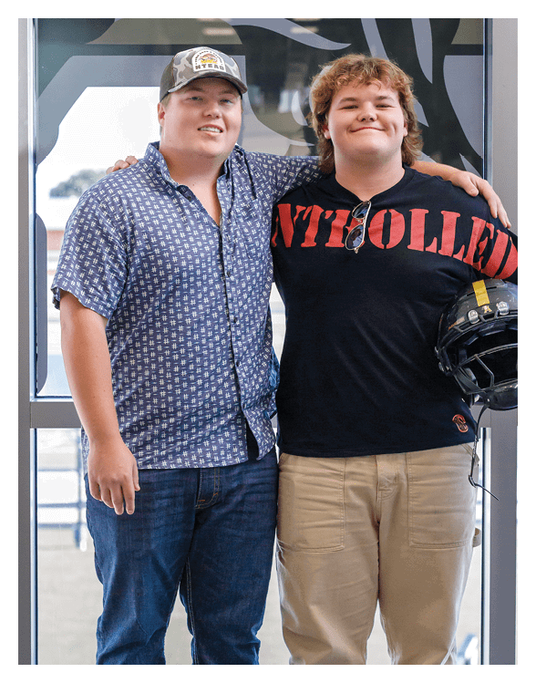 Two people stand close together with one person wearing a patterned blue button-up shirt and a cap while the other wears a black shirt with red lettering. Description generated by AI