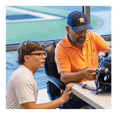 Two people wearing glasses are seated at a table with a black football helmet and a sticker, with one person wearing an orange shirt and a blue cap. Description generated by AI