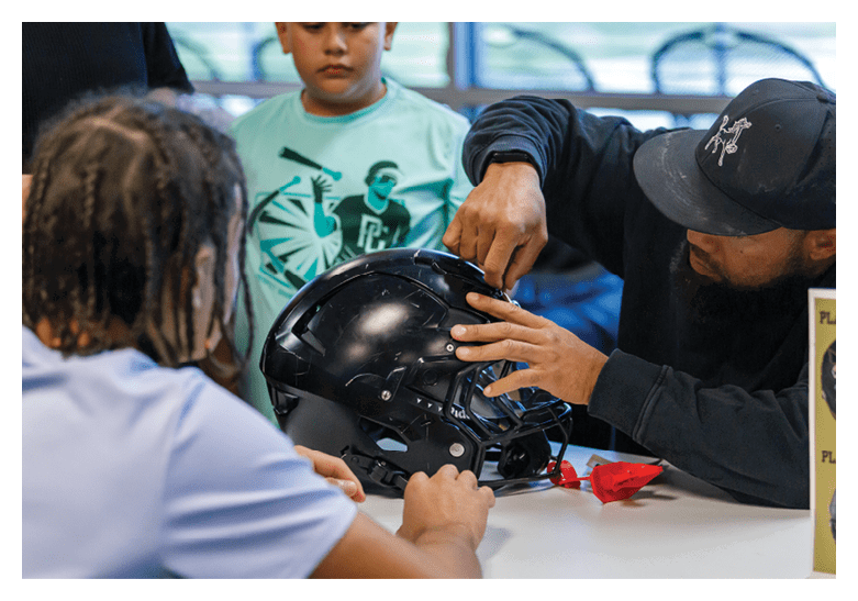 An adult wearing a black cap is adjusting a black football helmet on a table while two children watch attentively. Description generated by AI