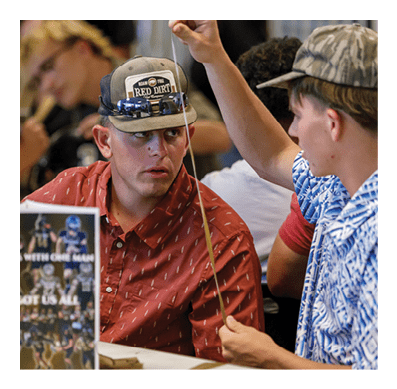Two adults are engaged in conversation at a table, one wearing a patterned red shirt and cap, and the other wearing a blue and white patterned shirt while holding a thin stick. Description generated by AI