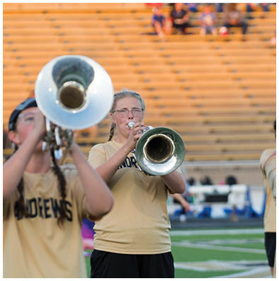 Two people wearing matching shirts are playing brass instruments in an outdoor setting with bleachers in the background. Description generated by AI