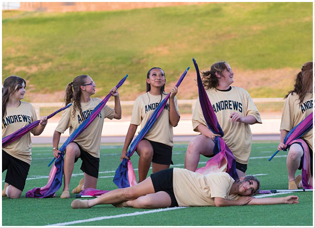 Four people wearing matching Andrews shirts and black shorts are kneeling on a field holding colorful flags, with one person lying down in front of them. Description generated by AI