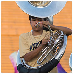 A person wearing a beige shirt with text is playing a large brass instrument, likely a sousaphone, against a blurred background with orange and purple colors. Description generated by AI