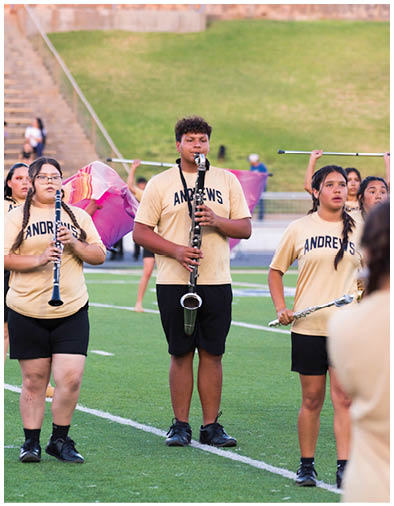 Person with curly hair wearing a light-colored shirt with the word “ANDREWS" playing a clarinet outdoors with a blurred green background. Description generated by AI