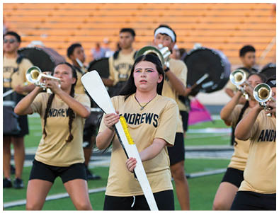 A group of people in matching uniforms are playing brass and percussion instruments in a stadium with empty orange bleachers in the background. Description generated by AI