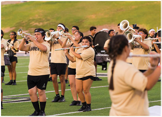 A group of people wearing matching beige shirts and black shorts are playing brass and woodwind instruments on a sports field. Description generated by AI
