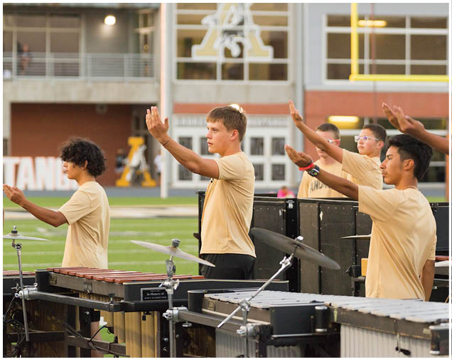 Several people wearing matching light-colored shirts are standing outdoors with their arms raised in a coordinated manner, appearing to perform or practice a routine near musical instruments. Description generated by AI