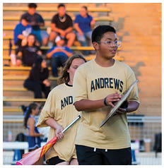 A person wearing glasses is in the foreground with blurred people sitting on bleachers in the background against a warm-toned wall. Description generated by AI
