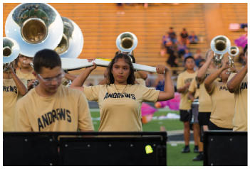 People in a marching band are playing brass instruments with one person holding a large white object above their head in an outdoor setting with bleachers in the background. Description generated by AI
