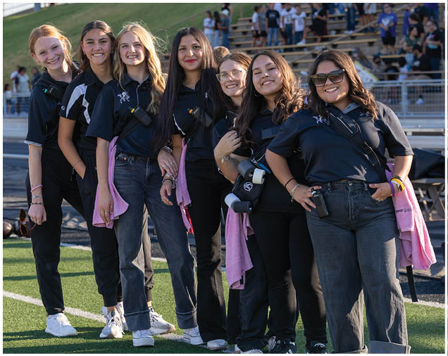 A group of five people wearing matching dark shirts and jeans stand closely together outdoors on a sports field, smiling and holding pink towels. Description generated by AI
