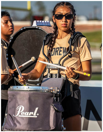 Person with braided hair wearing dark sunglasses and a beige shirt holding a lacrosse stick over their shoulder outdoors. Description generated by AI