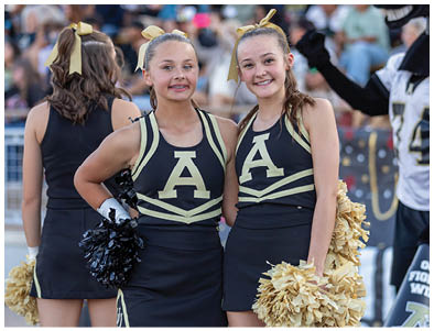 Two people wearing matching cheerleading uniforms with large letter A on the front smile while holding pom-poms at an outdoor event. Description generated by AI