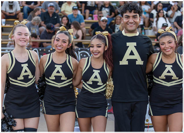 Five people wearing matching cheerleading uniforms with a large letter A on the front are standing closely together and smiling in front of a crowd in bleachers. Description generated by AI