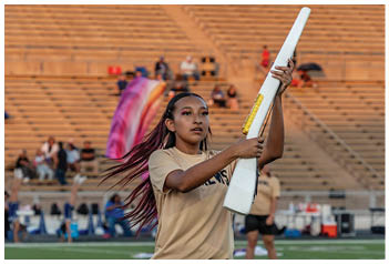 A person with long hair wearing a beige shirt and black shorts is holding a white and yellow object on a sports field with bleachers and other people in the background. Description generated by AI