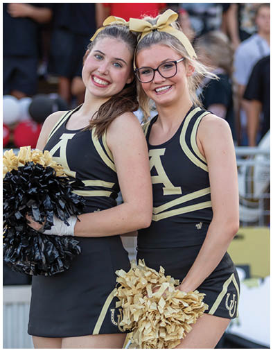 Two adults wearing matching black and gold cheerleading uniforms with bows in their hair smile while holding pom-poms and standing close together at an outdoor event. Description generated by AI