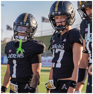 Three people wearing black football uniforms and helmets stand on a field with a blurred stadium background. Description generated by AI
