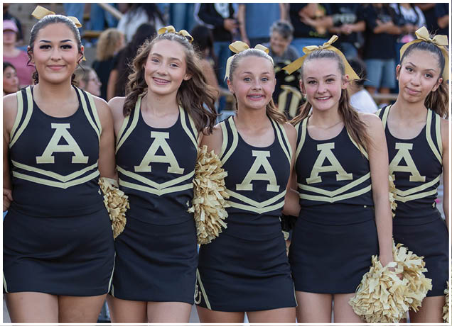 Five people wearing matching cheerleading uniforms with large letter A on the front stand closely together smiling, some holding gold pom-poms, with a crowd visible in the background. Description generated by AI
