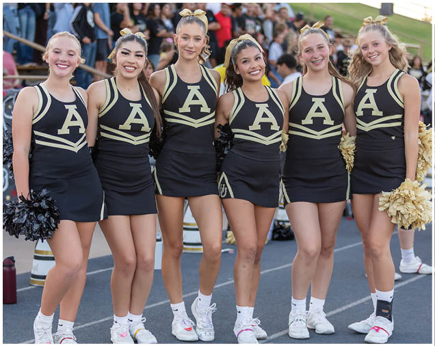 Six people wearing matching cheerleading uniforms with large letter A on the front stand side by side on a track with a crowd in the background. Description generated by AI