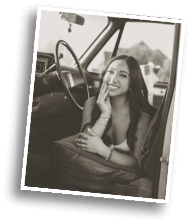 A person with long hair is smiling and resting their face on one hand while sitting inside a vintage car with a steering wheel visible. Description generated by AI