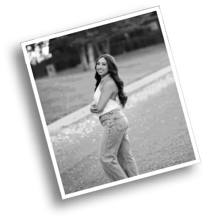 A person with long hair wearing a sleeveless top and jeans is standing outdoors on a path with trees in the background, smiling and looking over their shoulder. Description generated by AI