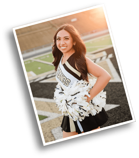 A person with long hair wearing a cheerleading uniform holding white and gold pom-poms stands on a sports field smiling. Description generated by AI