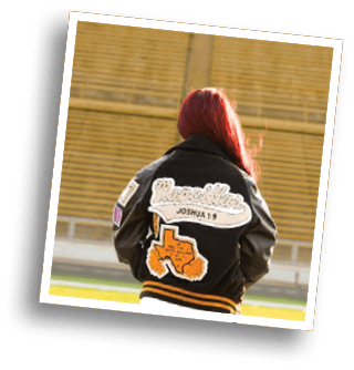 A close-up of a black garment with embroidered patches including the word “Mancil," a Bible verse reference "Joshua 1," and an orange Texas-shaped patch labeled "4A REG 1 DIV 2 STATE QUALIFIER." Description generated by AI