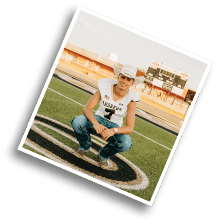 A person wearing a football jersey and jeans is crouching on a football field with a scoreboard and stadium seating in the background. Description generated by AI