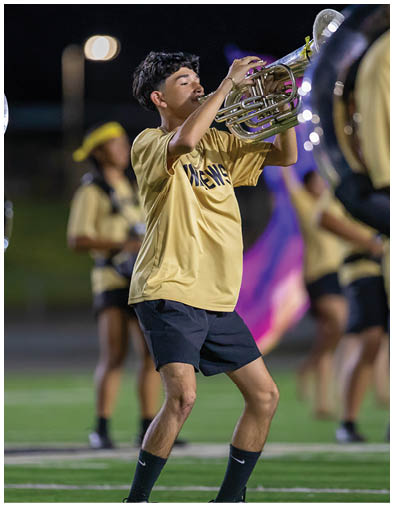A person wearing a yellow shirt and black shorts is playing a brass instrument on a field with other similarly dressed people in the background. Description generated by AI