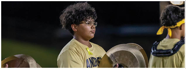 A person with curly hair and glasses is holding cymbals and wearing a yellow shirt while standing outdoors at night. Description generated by AI