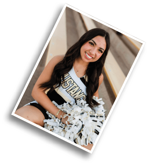 A person with long wavy hair wearing a cheerleading uniform that says “Mustangs" is holding white and gold pom-poms while sitting on a bench. Description generated by AI
