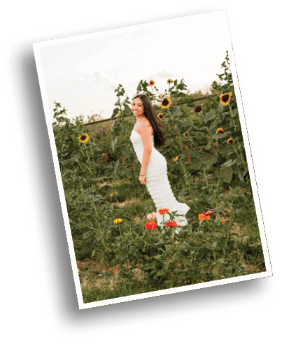 A person with long hair wearing a white dress is standing and smiling in a field of sunflowers and other wildflowers. Description generated by AI